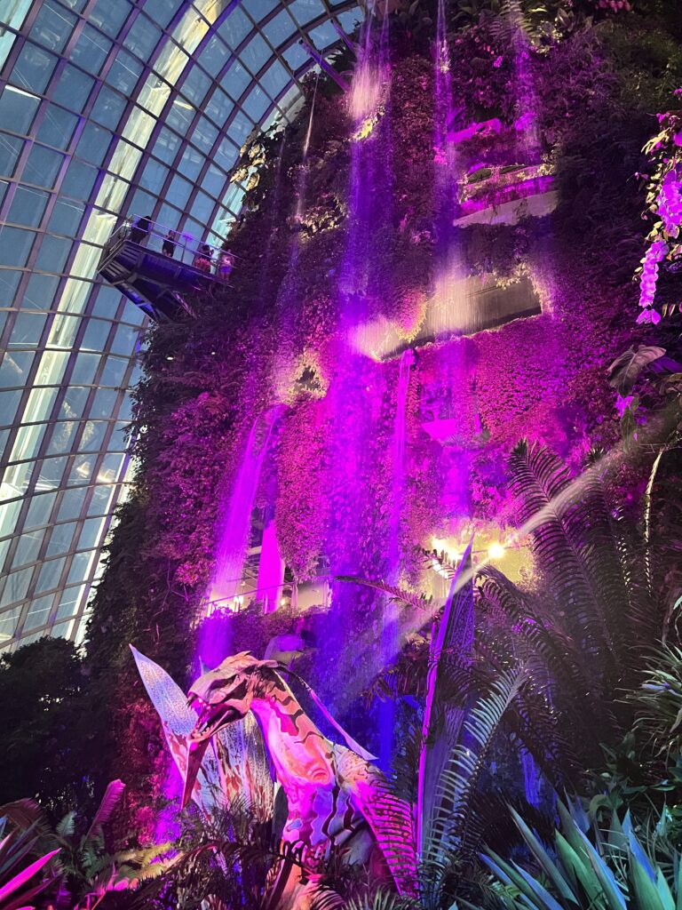 Cloud forest wall lit up with pink lights with one of the largest indoor waterfalls in the world with an Avatar character in front inside of the Gardens by the Bay in Singapore. 